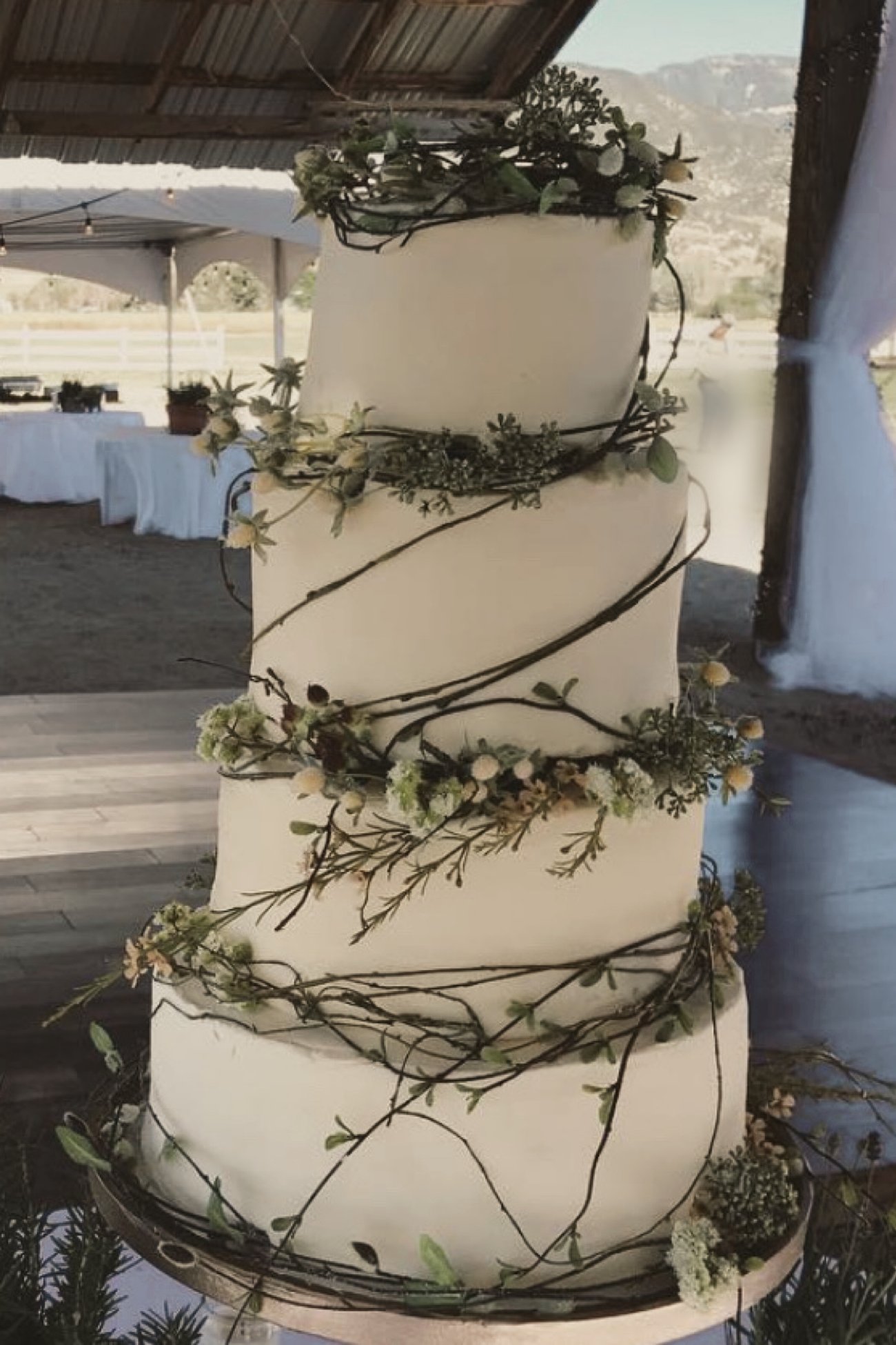 A four-tier white wedding cake wrapped in twigs and greenery, under a barn roof