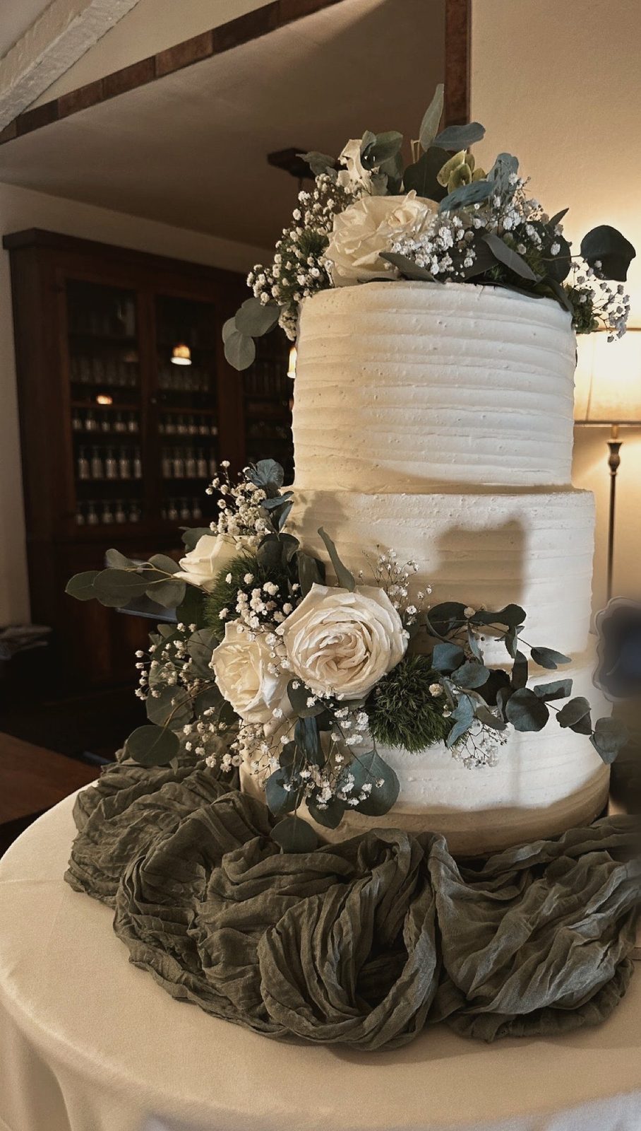 A three-tier textured white wedding cake with white roses, eucalyptus and baby’s breath on sage gauze