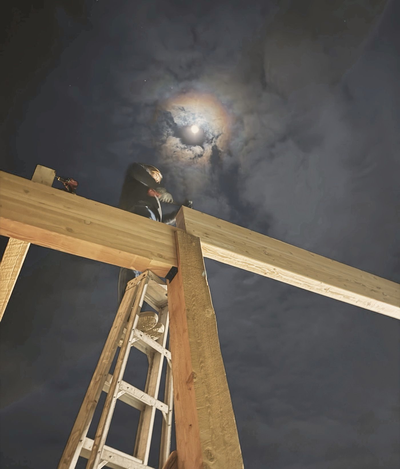 Looking up at the timber frame at night, a beam being set under a haloed moon