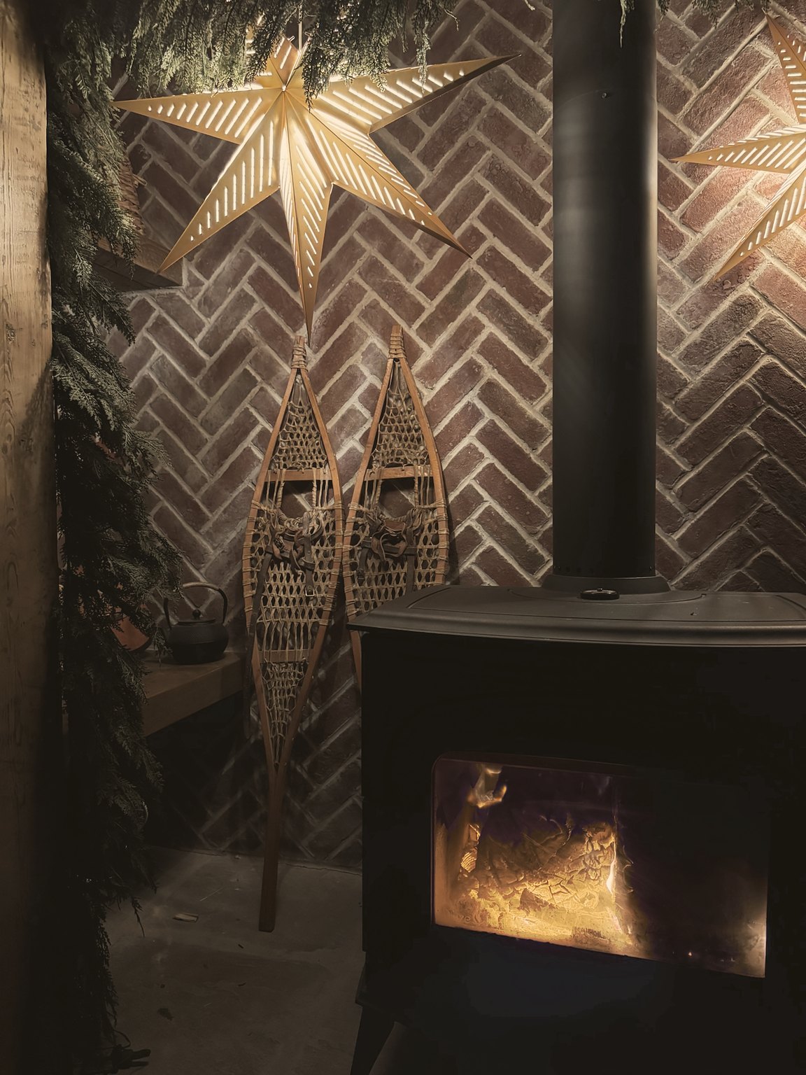 Glowing paper stars over vintage snowshoes on a herringbone brick wall, beside our black wood stove and cedar garland